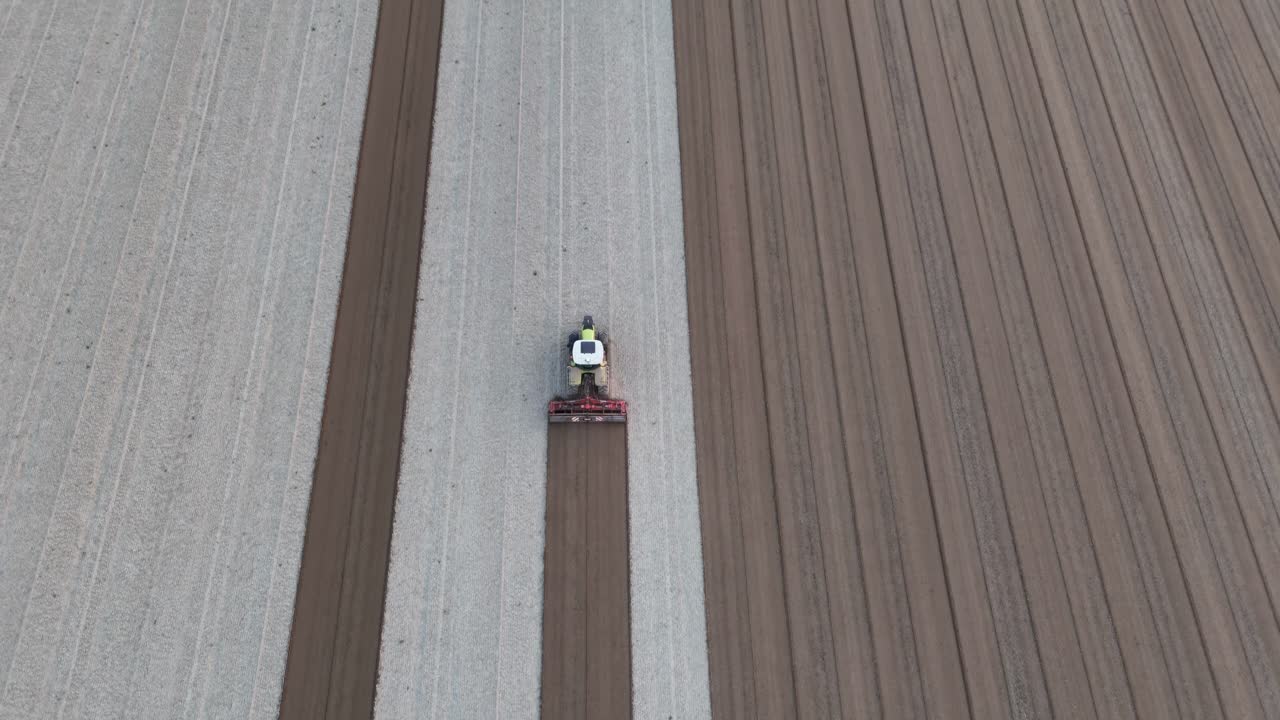 Tractor levels the field below, pulling harrow to form deep brown seed rows beside lighter furrows in vast, evenly plowed agricultural landscape under soft daylight, drone ascending shot, slow motion