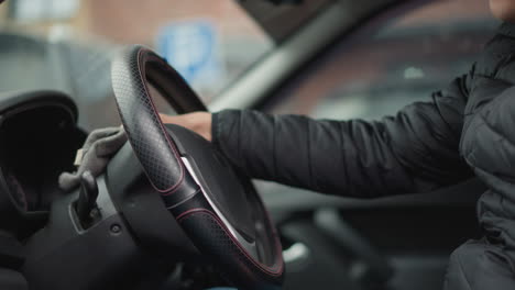 side view of urban explorer wiping leather steering wheel with towel in circular motion inside parked car cabin showing stitched wheel cover and blurred city background through window on cloudy day