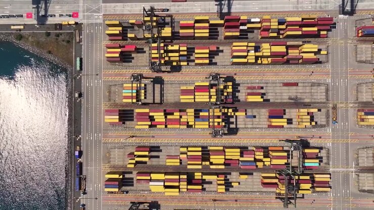 Container ships wait at the port due to shipping congestion. Cargo is stacked on docks, showing delays in freight transportation. Port operations continue despite challenges.