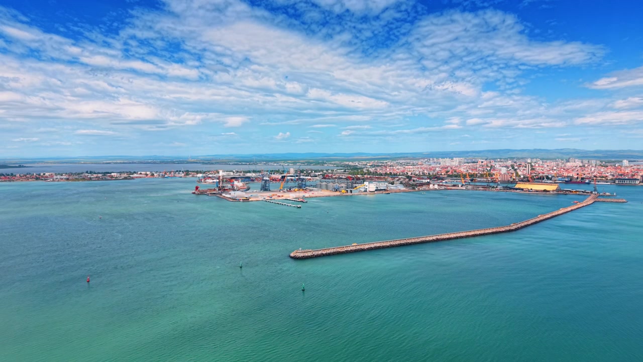 Terminale portuale industriale e molo frangiflutti. Ampia vista aerea di un lungo frangiflutti in pietra che protegge un porto industriale con gru e uno sfondo cittadino.