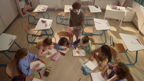 Maestro-De-Escuela-Explicando-La-Lección-A-Los-Estudiantes.-Niños-Inteligentes-Aprendiendo-En-Un-Escritorio-Redondo