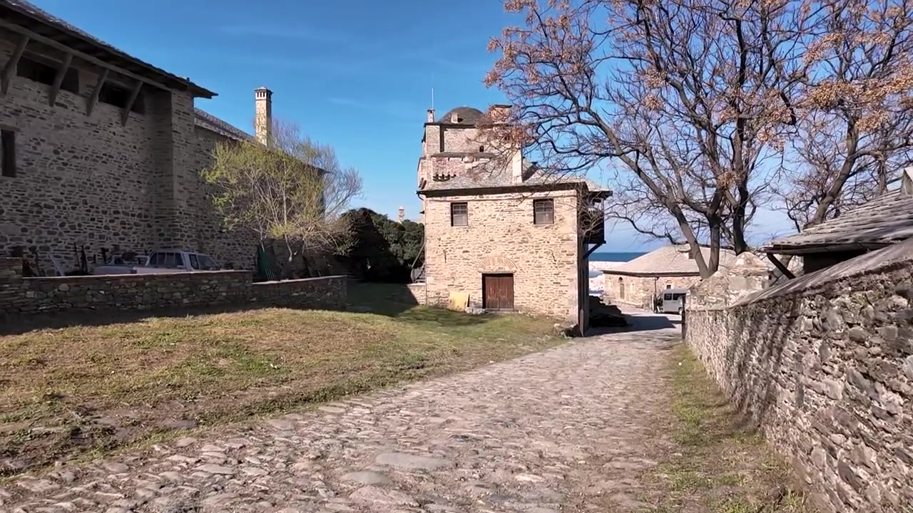 Un tour di un complesso monastico in Grecia. Il sole splende sugli edifici in pietra e sul sentiero acciottolato. I visitatori passeggiano e si godono le viste del paesaggio e dell'architettura.