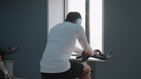 rear view of boy in white sportswear cycling on stationary bike with headphones on, listening to music, while another person sits and leans against gym wall under bright overhead lights