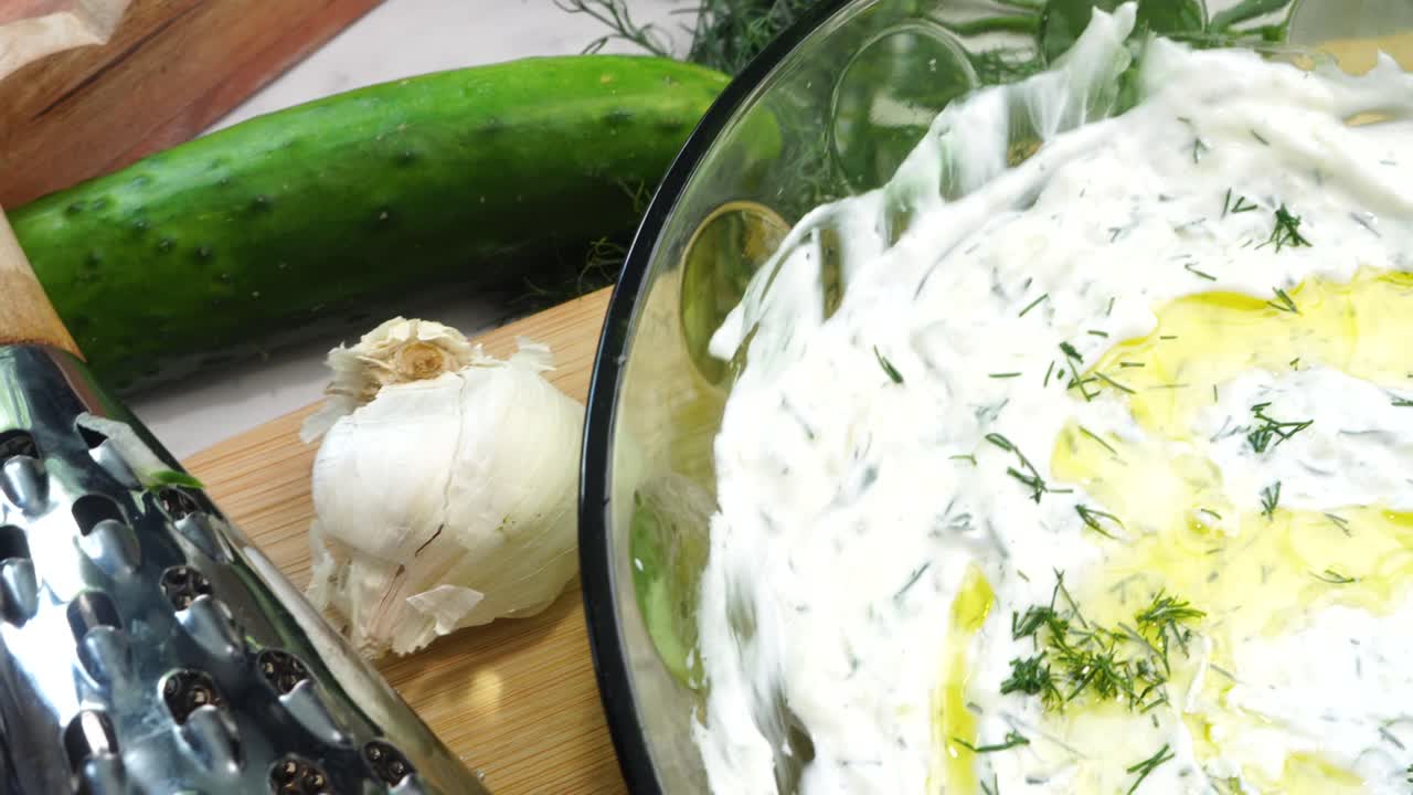 Making Tzatziki Greek or Mediterranean sauce or dip for a party. Dip or sauce in a green bowl on a wooden cutting board with dill, green napkin and a grater. Greek Tzatziki sauce for all foods.