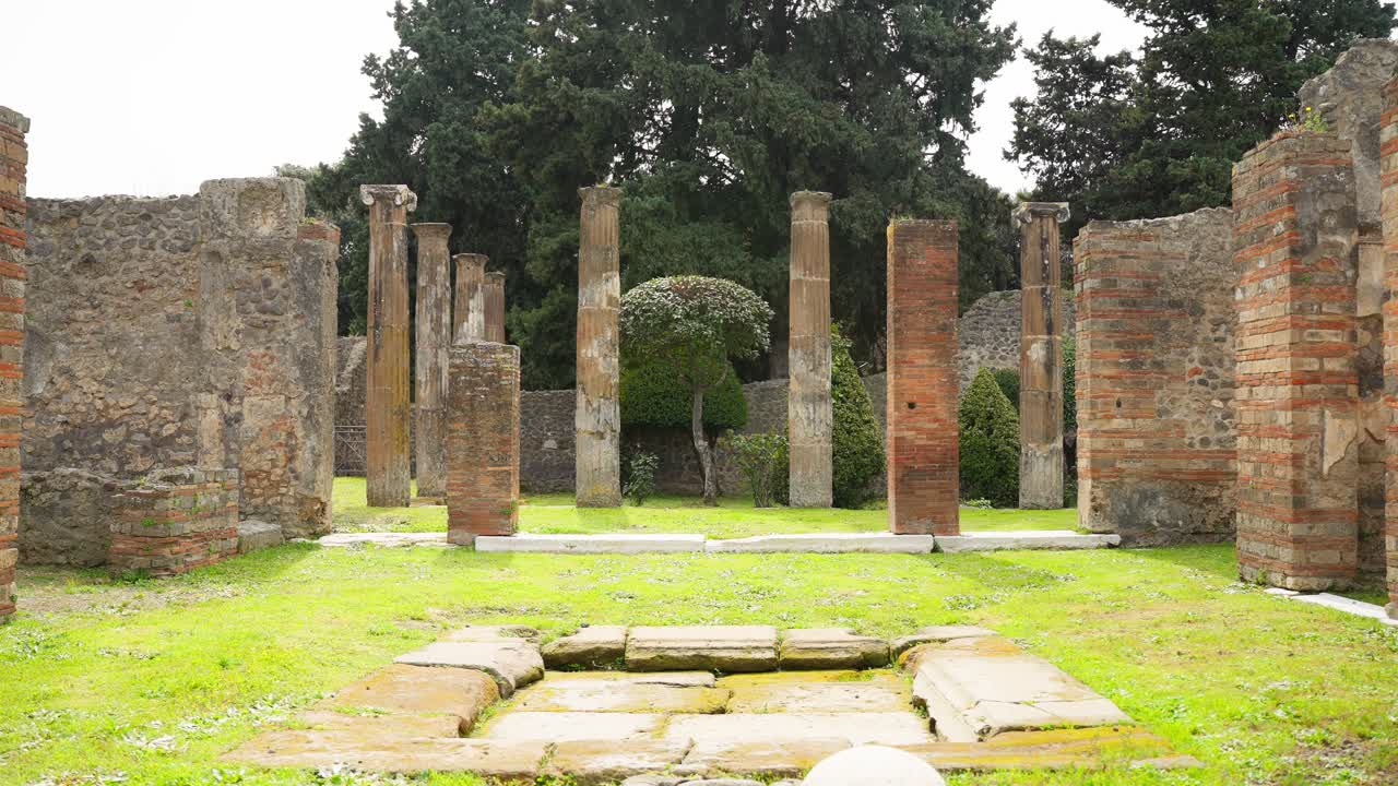 Ampia veduta di un cortile di Pompei con colonne erette, pilastri in mattoni e un elemento centrale in pietra, che cattura la disposizione a giardino aperto delle rovine dell'antica casa romana