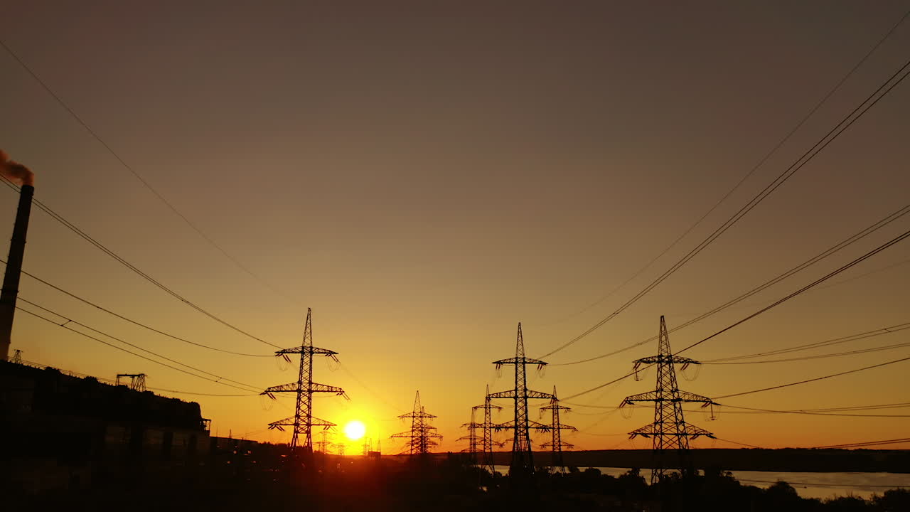 Sunset among high voltage towers. Transmission lines on the setting sun background. Electricity distribution in the evening. Motion camera back.