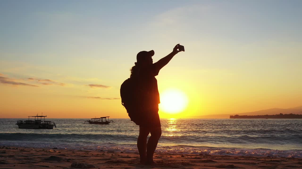 Girl taking photos with her smartphone on beautiful exotic beach washed by sea waves at sunset with dusty yellow sky over tropical bay