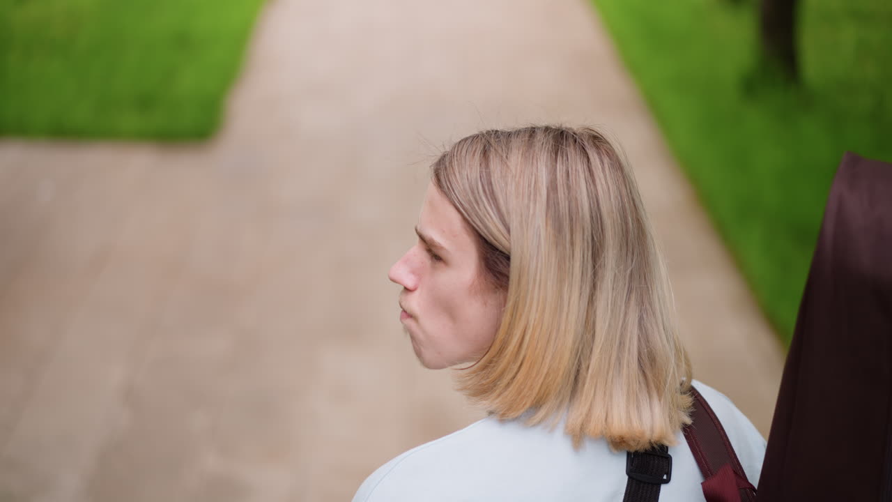 caucasian guitarist walking down park path with brown guitar case on back, overhead perspective, paved walkway flanked by grass, blonde hair, pastel sweater, steady stride, reflective mood, moving