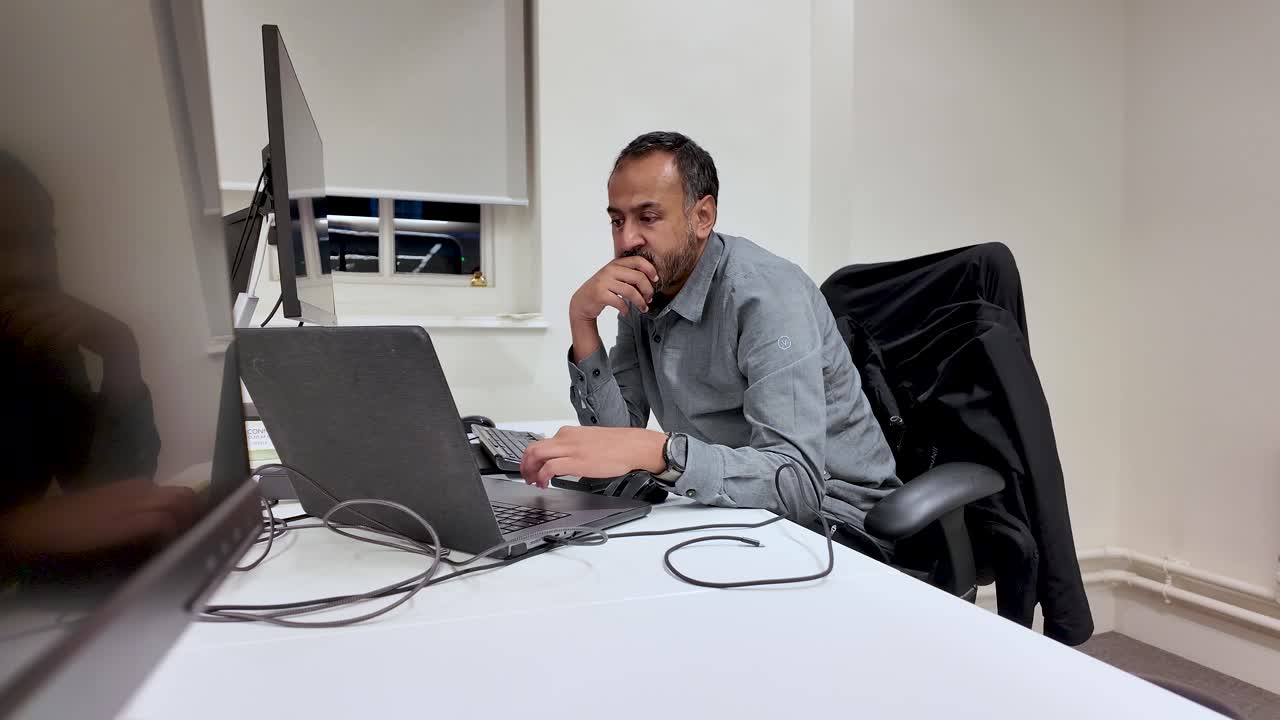 Südasiatischer Mann, der an einem Laptop an einem Bürotisch mit Mauspad und zweitem Monitor für Geschäfts- oder Technologiekonzepte arbeitet