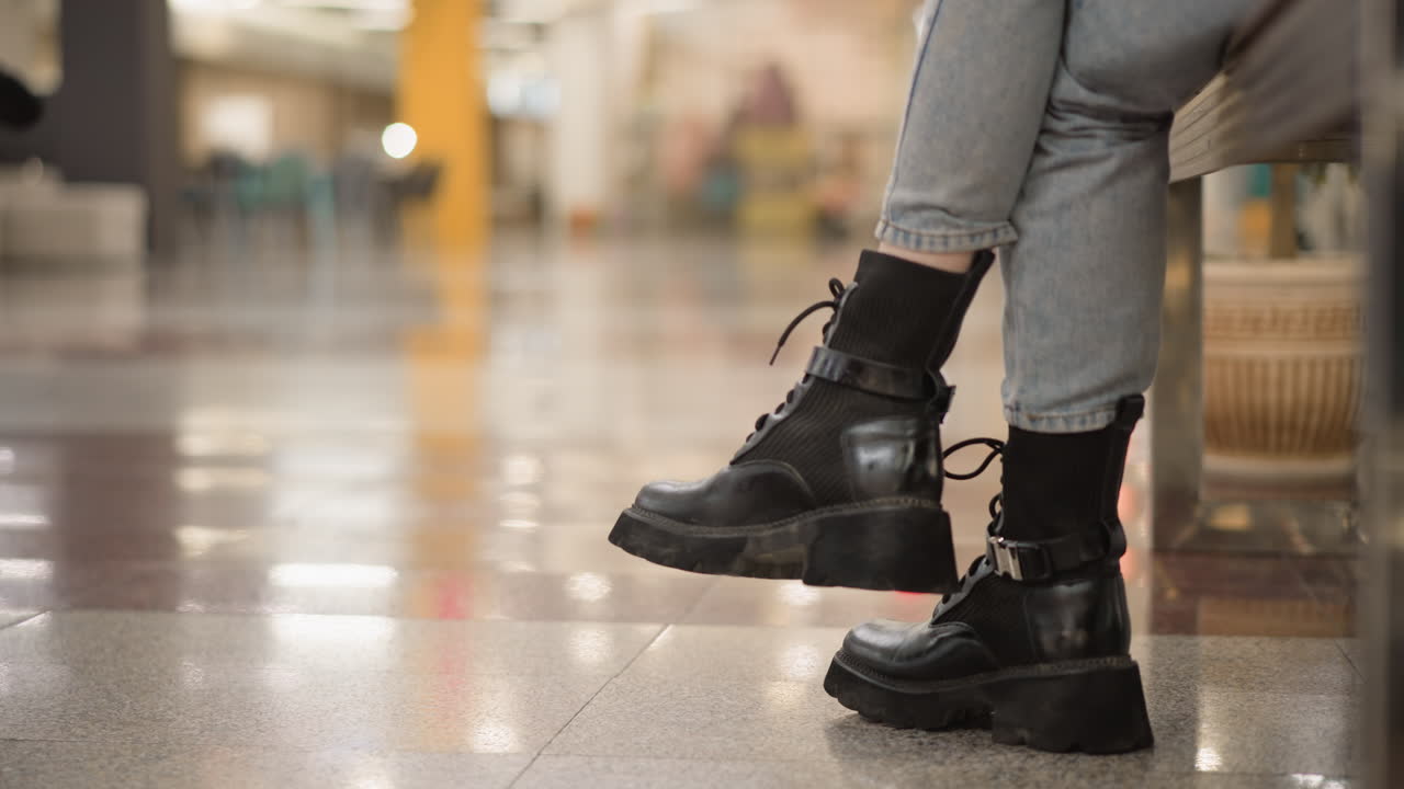leg view of poised shopper seated on mall bench with legs crossed wearing chunky black boots and denim jeans, background blurred people walking past under bright ceiling lights