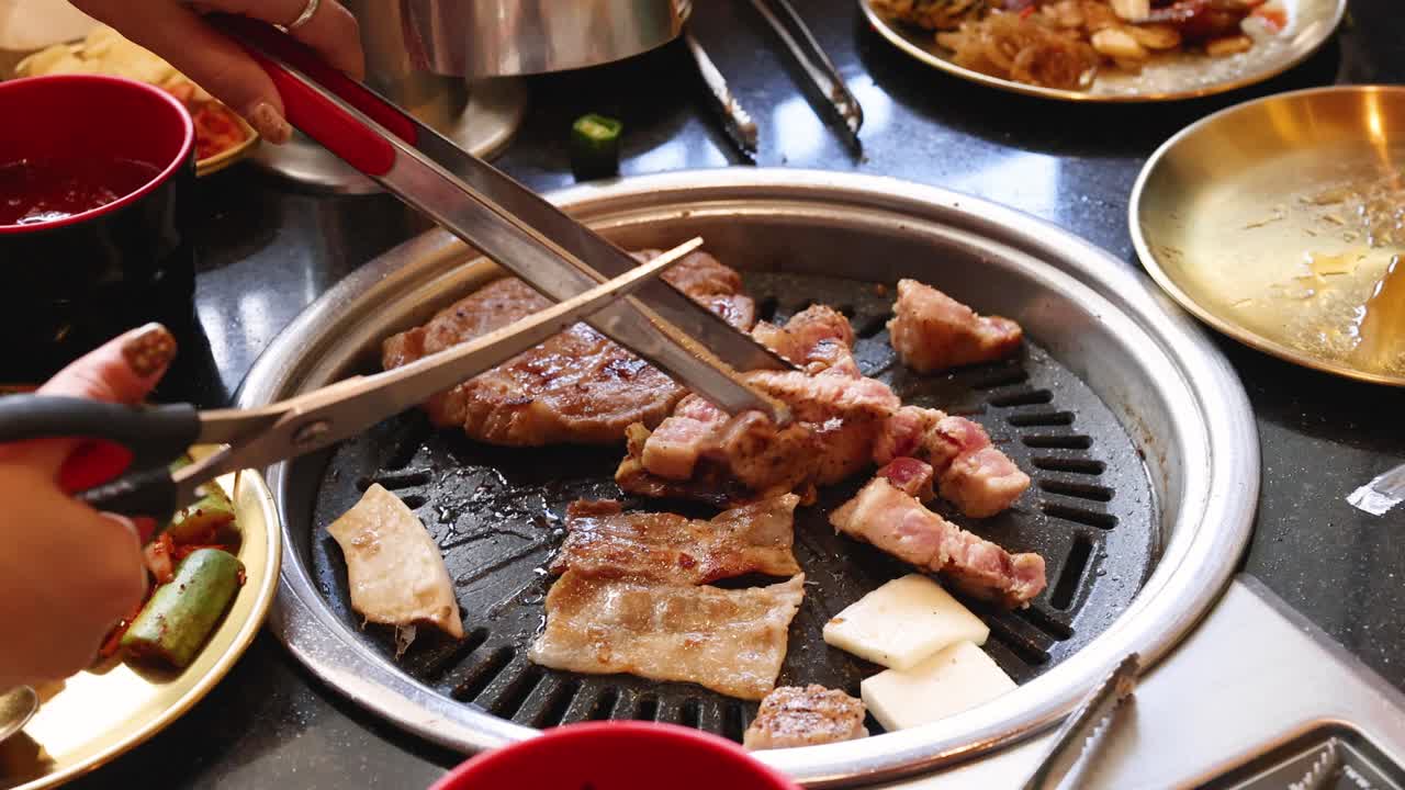 Hands slice grilled pork belly with scissors over tabletop grill, bright lighting, lively dining atmosphere
