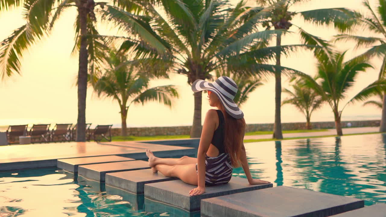 Sexy long hair brunette in monokini sits on stone in pool at golden sunset