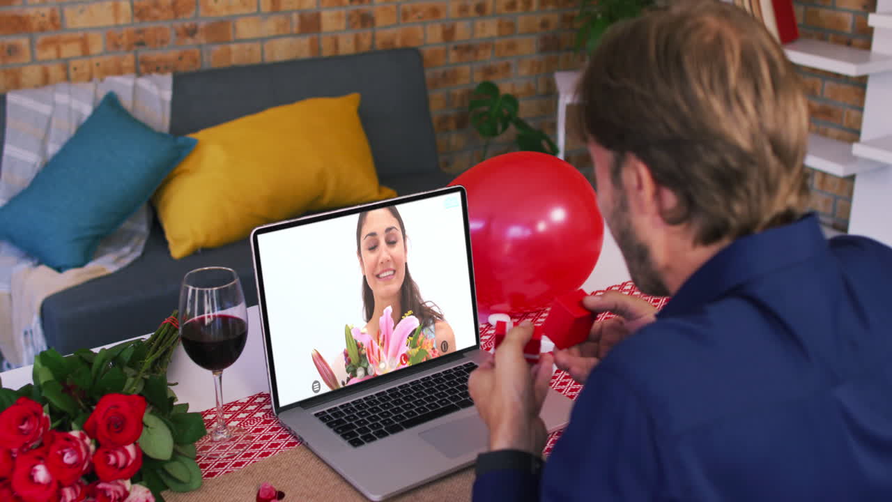 Casal caucasiano em videochamada de Dia dos Namorados, homem mostrando anel para mulher sorrindo na tela do laptop.