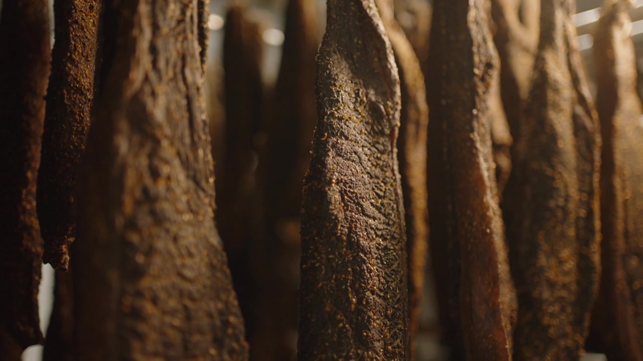 Spesse fette di biltong essiccato pendono da binari metallici in una struttura di stagionatura sudafricana, con superfici dalla texture ruvida in luce calda.