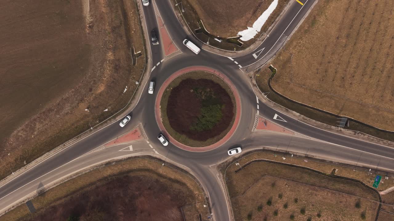 Top-down aerial drone view of a roundabout traffic intersection with several cars circulating on the road. The circular junction connects multiple rural roads and is surrounded by agricultural fields