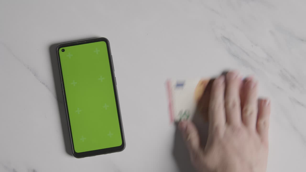 fotografía aérea de una mano agarrando un billete de 10 euros junto a un teléfono móvil de pantalla verde