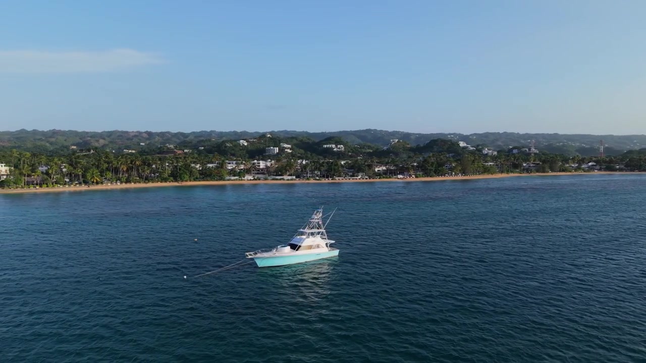 Droneoptagelse fra luften, der kredser om en forankret sportsfiskeri-yacht i en rolig bugt, skiftende fra et lyst modlys fra solnedgangen for at afsløre den tropiske kystlinje ved Las Terrenas.