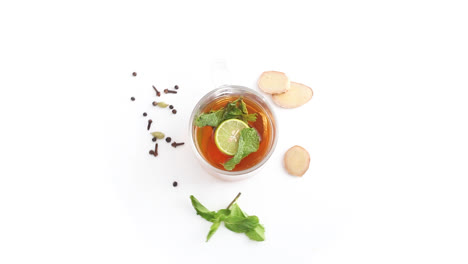 Té-De-Limón-En-Una-Taza-De-Vidrio-Transparente-Con-Rodajas-De-Limón-Y-Hojas-De-Menta-De-Jengibre-Clavo-De-Olor-Cardamomo-Pimienta-Negra-En-Fondo-Blanco-Tiro-Superior-Giratorio