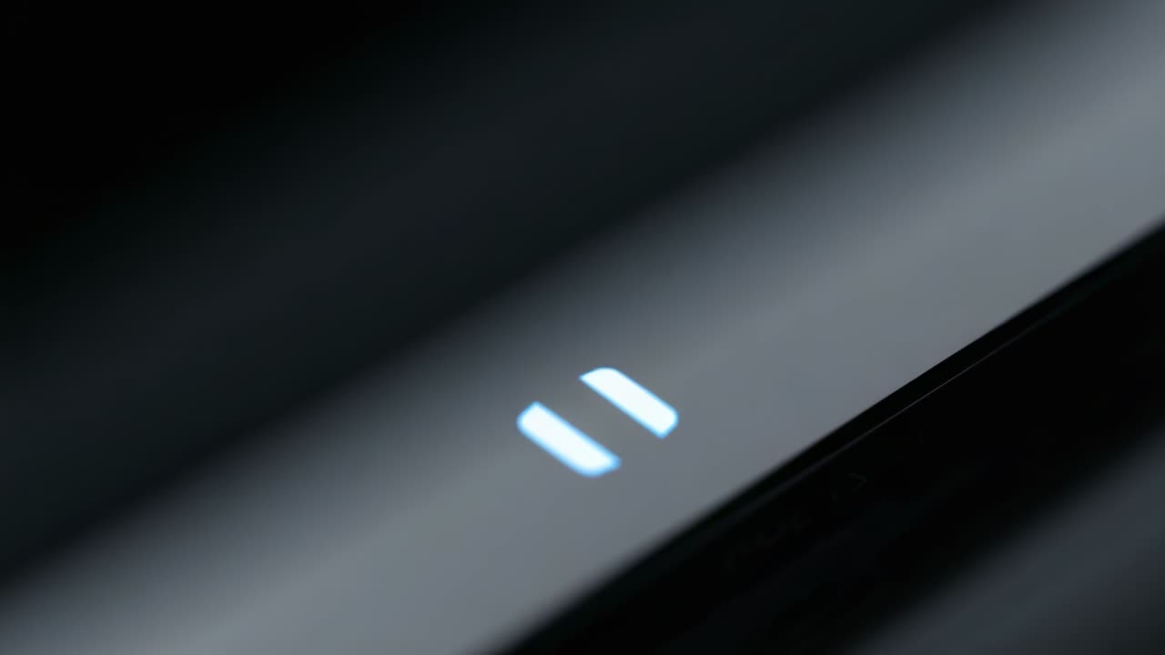 Camera tightening on glossy black tech device, pause icon brightening, highlighting blue LED detail