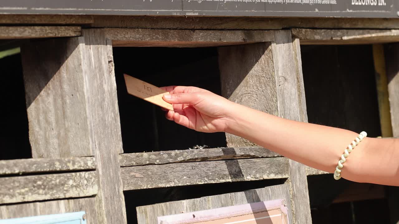 A person’s hand, wearing a bracelet, throws a piece of paper into a rustic wooden trash bin under bright natural sunlight. Static camera, clear shadows