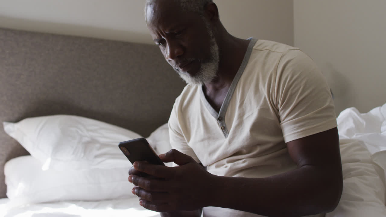 African american senior man using smartphone while sitting on the bed at home