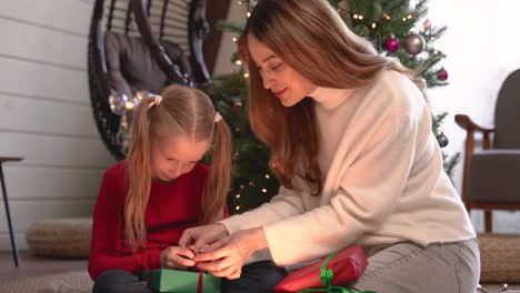 mother and daughter wrapping christmas presents sitting on the floor near a christmas tree in living room