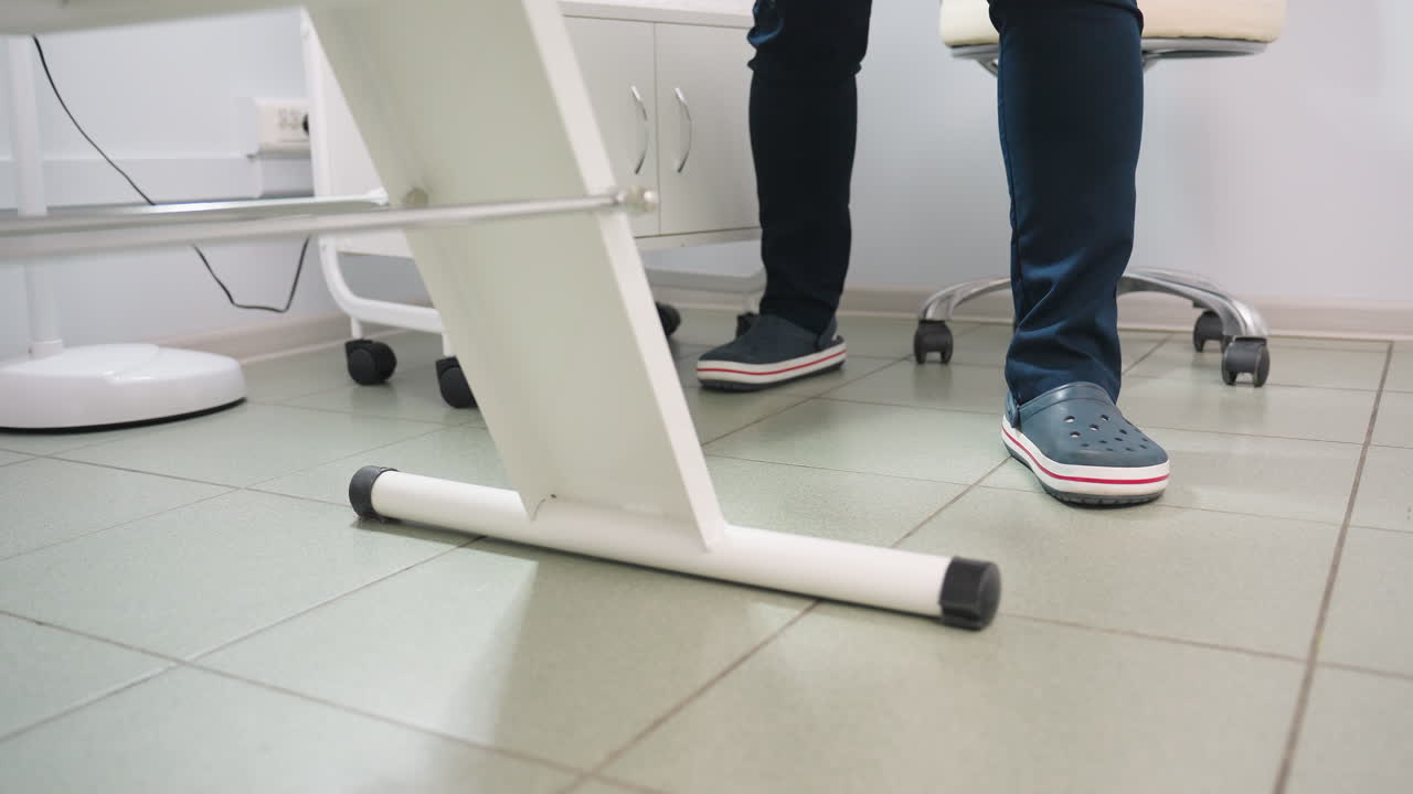 Leg view of practitioner on crocs wearing navy blue uniform standing beside clinical bed under bright lamp with chair and product table visible in bright treatment room with tiled floor