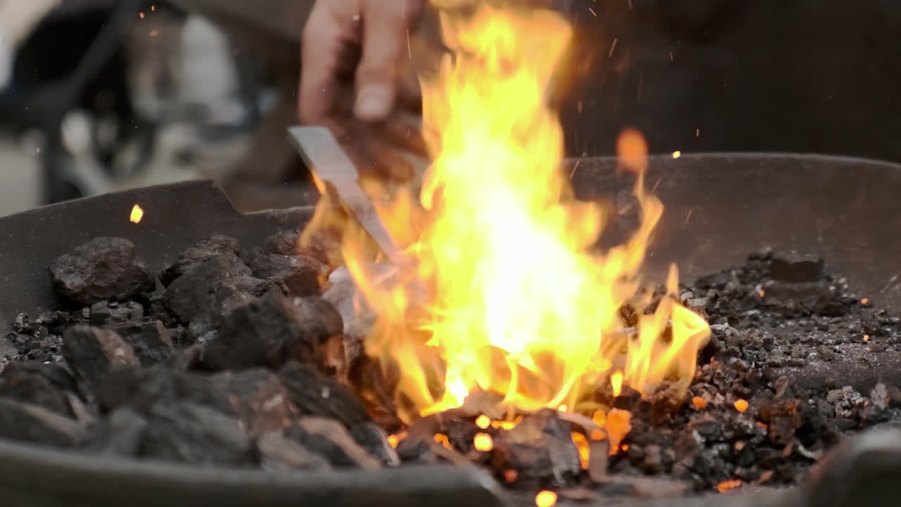 Super slow-motion static close-up as a blacksmith feeds a metal piece into a coal forge; bright flames and flying embers in the foreground, gritty workshop atmosphere