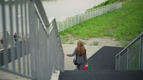 back view of lady holding colorful shopping bags while walking down stairs, holding railing for support, with three individuals in the background, featuring a river, lamp post, and greenery