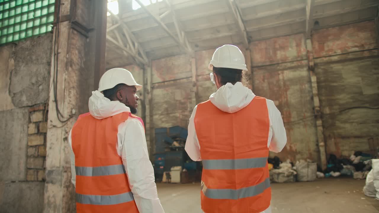 Rear view of two male colleagues in white uniforms and orange vests going out into the premises of a huge waste recycling and sorting plant. A Black man and a brunette man with a beard are talking and walking around a large plant