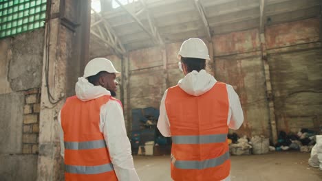 Rear-view-of-two-male-colleagues-in-white-uniforms-and-orange-vests-going-out-into-the-premises-of-a-huge-waste-recycling-and-sorting-plant.-A-Black-man-and-a-brunette-man-with-a-beard-are-talking-and-walking-around-a-large-plant