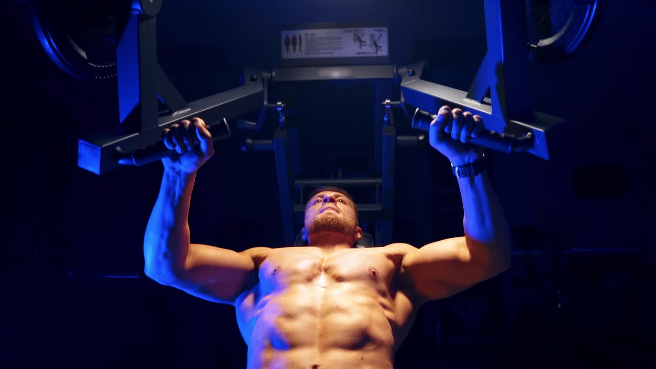 Handsome young man doing bench press workout in gym. Blue light filter. Strong abs. Heavy lift. Video from below.