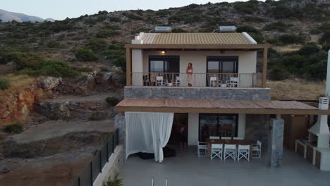 mujer de cabello oscuro está de pie en la terraza de una casa y disfruta de la vista - avión no tripulado volando hacia atrás al amanecer en grecia en la isla de creta - se pueden ver palmeras y montañas rocosas