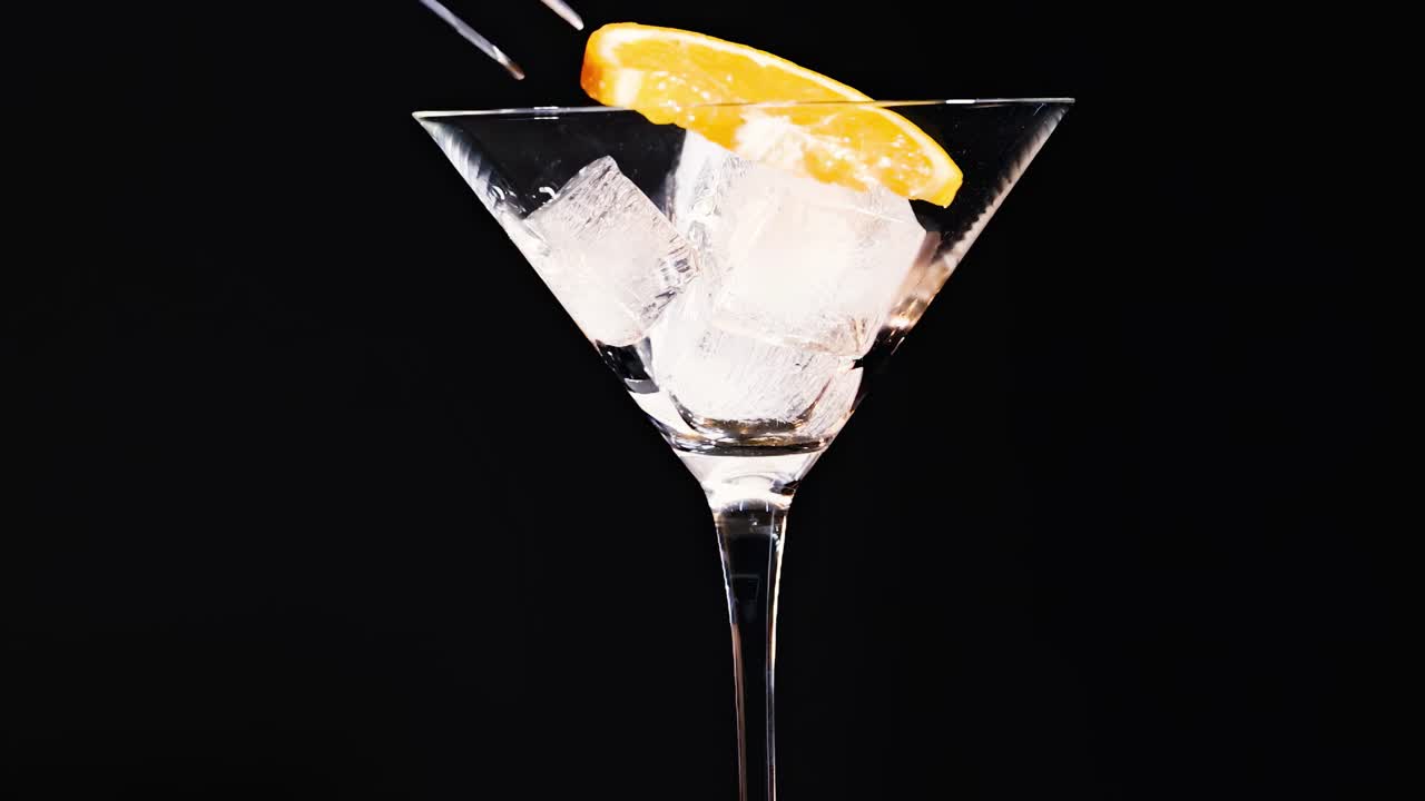A martini glass filled with ice cubes and garnished with an orange slice against a black background.