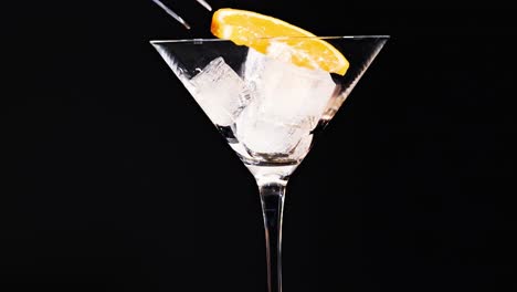 a martini glass filled with ice cubes and garnished with an orange slice against a black background.