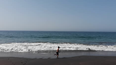 Aerial side view man running along black sandy beach and seashore during sunny day, Gui Gui Beach, las Palmas de Gran Canaria