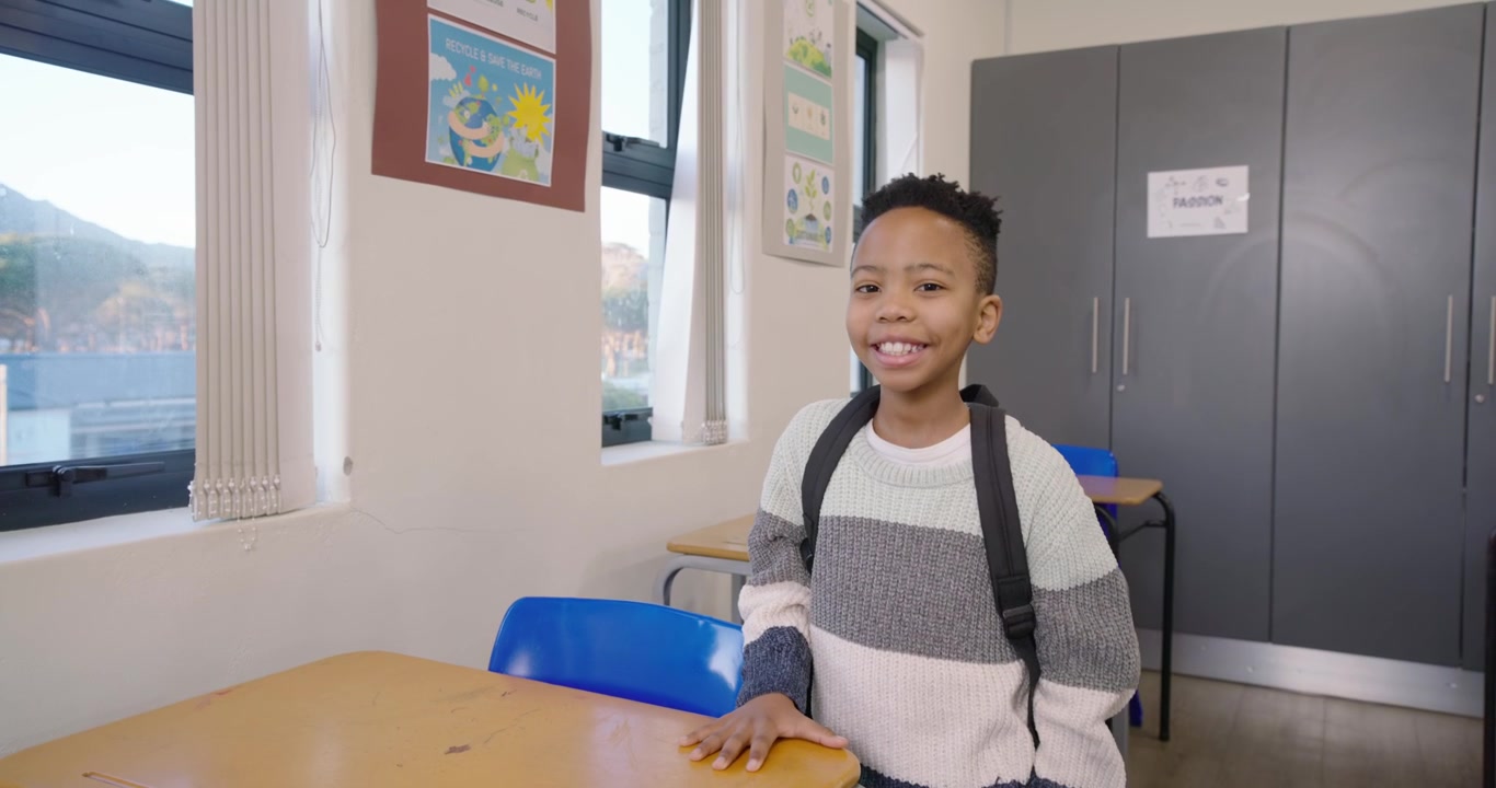 Bambino africano in piedi accanto alla scrivania vicino alle finestre, con la telecamera che si avvicina, che si gira sorridendo con lo zaino, con spazio per il testo.