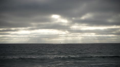 La-Vela-De-Un-Velero-Distante-Se-Ve-En-El-Horizonte-Del-Océano-En-Un-Día-Sombrío-Con-Rayos-De-Sol-Brillando-A-Través-De-Las-Nubes