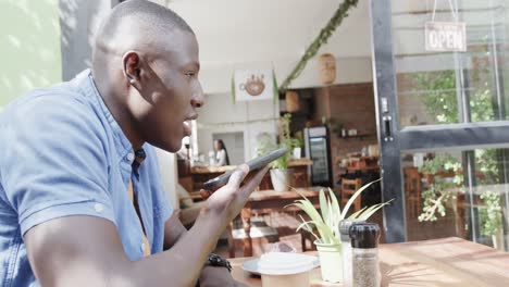 Happy-african-american-man-talking-on-smartphone-at-table-outside-coffee-shop,-slow-motion