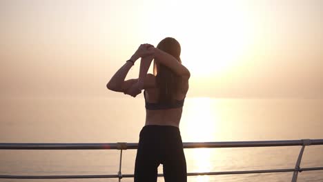 vista trasera de una niña delgada de cabello largo mirando al amanecer cerca del agua. estira los brazos. leggins y sujetador deportivo. enorme flexibilidad