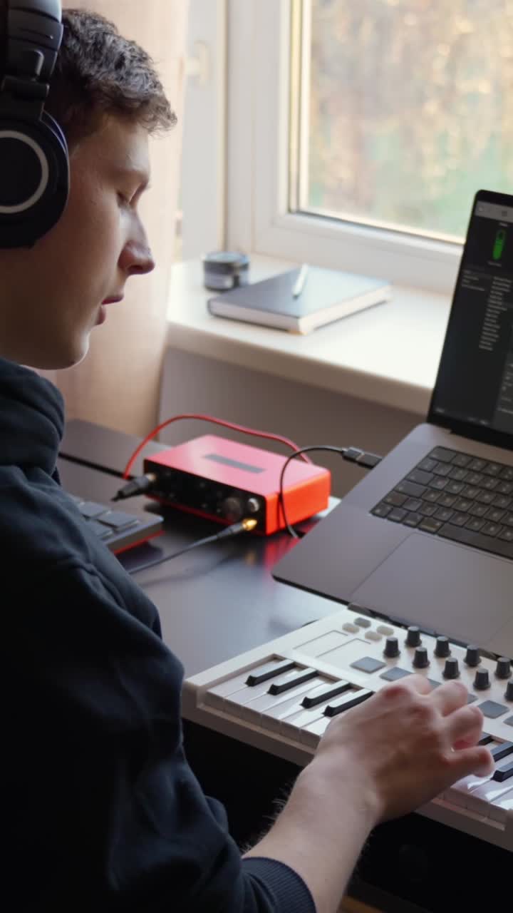 Vertical close-up from behind of a musician using a MIDI keyboard while wearing headphones in a home studio