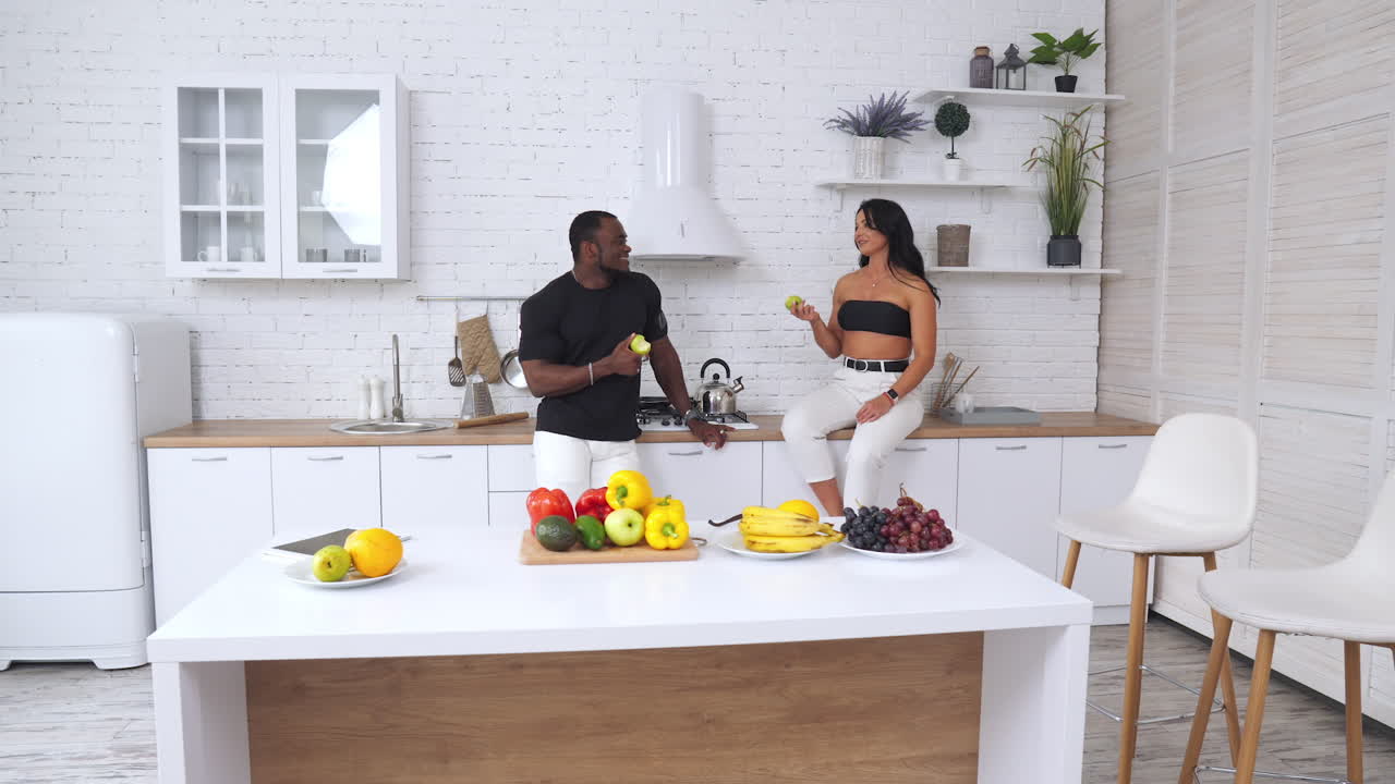 Healthy and fit couple having conversation and eating apples in the kitchen. Cheerful people talking near the table full of healthy organic food.