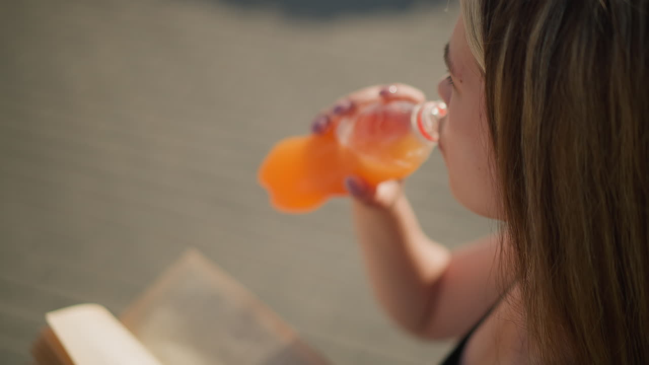 밖에서 앉아서 한 손으로 책을 들고 다른 손으로 병에서 주스를 마시고 있는 여성의 클로즈업, 책이 약간 흐릿하고, 그녀는 배경도 흐릿하게 읽고 있습니다.