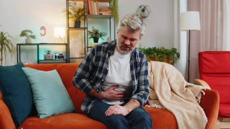 mature middle-aged man sits on couch feeling sudden strong abdominal stomach ache gastritis problem