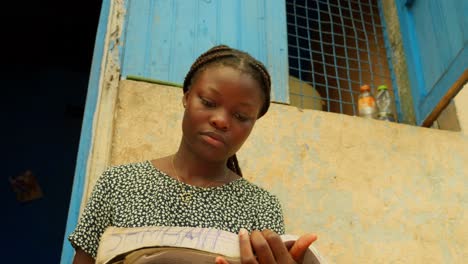Durch-Das-Lesen-Eines-Buches-Lernt-Ein-Mädchen-Außerhalb-Ihres-Zuhauses-In-Einem-Dorf-In-Kumasi,-Ghana,-Eine-Grundausbildung-Im-Lesen-Und-Schreiben