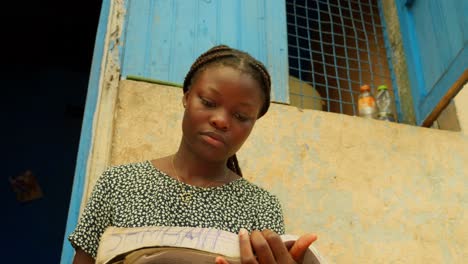 Al-Estudiar-Leyendo-Un-Libro,-Una-Niña-Está-Obteniendo-Educación-Básica-En-Lectura-Y-Escritura-Fuera-De-Su-Casa-En-Una-Aldea-De-Kumasi,-Ghana,-África