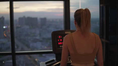 Woman-walking-on-treadmill-at-the-gym-with-cityscape-view-on-a-sunset-through-the-glass-window,-static-closeup
