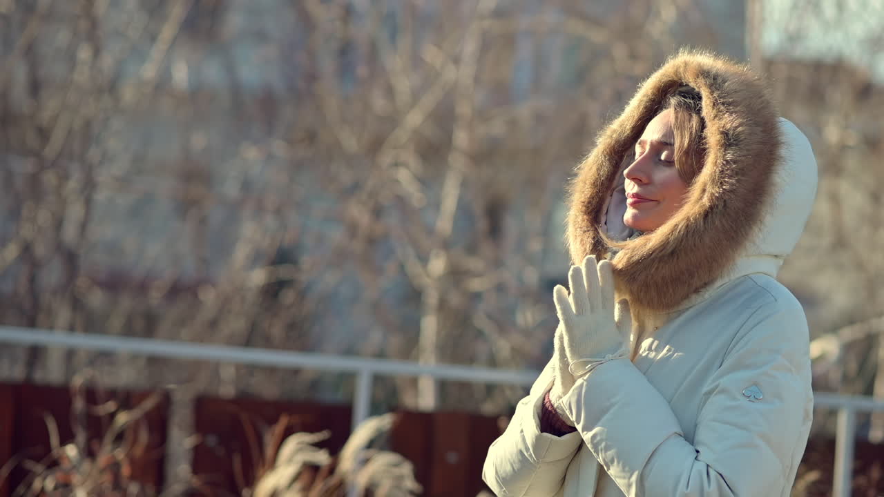 A warm, sunny winter day shows a woman standing outside in a hooded jacket. She smiles while adjusting her gloves, enjoying the bright weather and fresh air