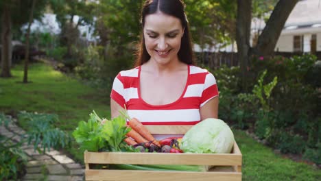 mujer caucásica sonriente de pie en el jardín sosteniendo una caja de verduras