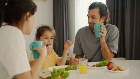 A-brunette-man-in-a-gray-T-shirt-holds-a-turquoise-cup-in-his-hand-and-has-breakfast-with-his-wife-and-little-daughter-in-a-yellow-dress-with-a-white-table-in-a-modern-kitchen